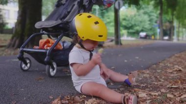Sarı miğferinde kuru yaprak, arka planda bebek arabasıyla yerde oturan çocuk, sonbahar öğleden sonra ağaçların olduğu parkta rahat bir şekilde oturuyordu.