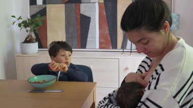 Anne yeni doğan bebeği emzirirken, büyük oğul yemek masasında oturuyor, yemek saatinde dikkati dağınık görünüyor, iki küçük çocuğu yetiştirmenin günlük hokkabazlığını temsil ediyor.