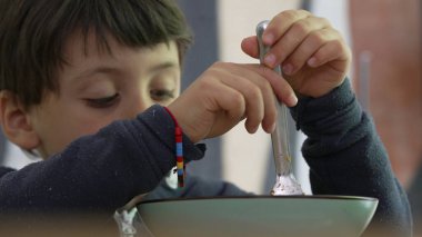 Çocuk elinde çatalla spagetti çeviriyor, işine derinden konsantre oluyor, kendini besleme sürecini ve tanıdık bir ev ortamındaki odaklanmış çabayı vurguluyor.