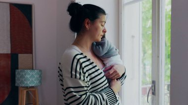 Anne pencerenin kenarında durmuş, yeni doğmuş bir bebek omuzunda, dışarıya bakıyor. Sahne, anne ve bebek arasındaki sakinlik, bağ ve huzur dolu anları aktarır.