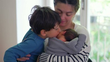 Büyük oğlan hem anne hem de yeni doğmuş bebeği öpüyor, sevgi ve bağla dolu şefkatli ve yürek ısıtan bir aile anı yaratıyor, anne ve kardeş arasındaki bağı vurguluyor.