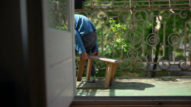 Genç bir çocuk balkonda küçük bir taburede oturuyor, bacaklarını açmış, dışarıda huzurlu bir anın tadını çıkarıyor. Güneşin tadını çıkaran bir çocuğun rahat ve kaygısız havası.