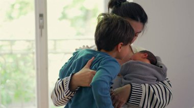 Anne hem yeni doğan hem de büyük çocuğu kucaklıyor, hassas bir aile anında kardeşler arasındaki sevgi ve ilgiyi dengeliyor, anne olmanın önemini vurguluyor..