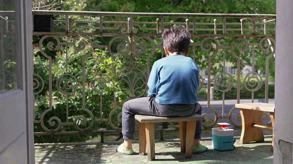 Genç bir çocuk balkonda küçük bir taburede oturuyor, yüzünü çevirmiş, dışarıda sakin bir anın tadını çıkarıyor. Doğayla iç içe olan bir çocuğun huzur dolu yalnızlığı..