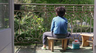 Genç bir çocuk balkonda küçük bir taburede oturuyor, yüzünü çevirmiş, dışarıda sakin bir anın tadını çıkarıyor. Doğayla iç içe olan bir çocuğun huzur dolu yalnızlığı.