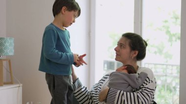 Küçük çocuk, bir bebeği kucağında tutarken annesine sevgiyle sarılıyor. Ailevi bir sevgi ve bağ anı yaratıyor. Evdeki sıcaklığı, şefkati ve güçlü aile bağlarını somutlaştırır.