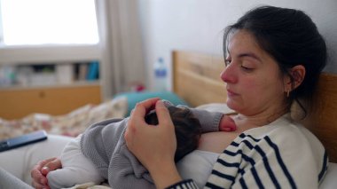 Anne, uyuyan bebeği göğsünde yatağında dinlenirken, gözleri bir an için sessiz bir yansımayla kapanıyor. Anne ve bebek arasında paylaşılan huzur ve huzur..