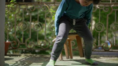 Genç bir çocuk bir tabureyi sürüklüyor ve balkona oturup tek başına oynuyor. Çocuk çevresini keşfederken bağımsız bir oyun ve sessiz bir keyif anı..