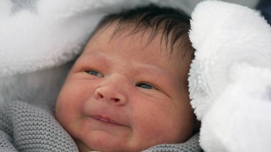 Yeni doğmuş bebek rahat bir battaniyeye sarılmış hafif bir sırıtışla, içerik gösteren ama biraz da eğlendirici bir ifade, erken yaşamda sıcak ve güvenli bir anı yakalıyor..
