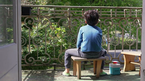 Genç bir çocuk balkonda küçük bir taburede oturuyor, yüzünü çevirmiş, dışarıda sakin bir anın tadını çıkarıyor. Doğayla iç içe olan bir çocuğun huzur dolu yalnızlığı.