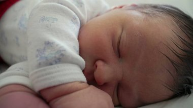 Yeni doğmuş bir bebeğin yüzü ve eline odaklanmış bir şekilde uyuduğu yakın plan bebeklik döneminin yumuşak, huzurlu ve huzurlu atmosferini yakalıyor.