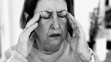 Şiddetli baş ağrısı ile mücadele eden yaşlı bir kadın, gözle görülür konsantrasyonda şakaklara masaj yapıyor. Yüz hatları ve yüz ifadeleri yoğun acıyı, zihinsel gerginliği, duygusal sıkıntıyı yansıtır.