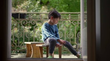 Genç çocuk bir balkonda ahşap bir taburede oturuyor, düşünceli bir şekilde aşağı bakıyor, çevrende yeşil bir ortam, ağaçların arasından süzülen doğal ışık - SD 480p