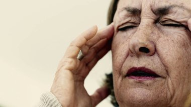 Yaşlı bir kadın şakaklarına sıkıca masaj yapıyor. Acıyı veya stresi azaltmaya odaklanıyor. Derin kırışıklıklar, yoğun yüz ifadesi, görülebilir gerilim şiddetli baş ağrısı gösteriyor. SD 480p