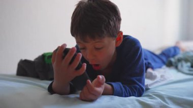 Küçük çocuk elinde akıllı telefonla yatakta uzanıyor, video izlerken ya da oyun oynarken odaklanmış ifade ediyor, evde film izlerken huzurlu ve sessiz bir çocukluk geçiriyor.
