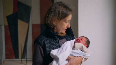 Büyükanne sevgiyle kundaklanmış yeni doğmuş bir bebeği kollarında tutuyor. Büyükannem, bebekle ilgilenirken şefkatle aşağı bakar. Bir anlık bağ ve özen gösterir..