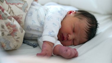 Yeni doğmuş bebek, yumuşak tulum giymiş bir yatakta huzur içinde uyuyor. Yavrunun sakin ifadesi ve rahat duruşu rahat bir ortamda huzurlu bir uyku anı yakalar.