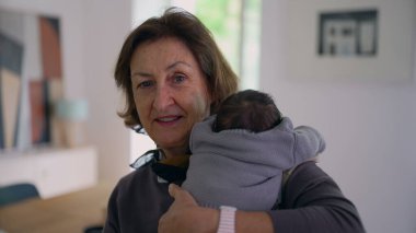 Büyükanne yeni doğmuş bir bebeği omzunda tutuyor, kameraya sıcak bir şekilde gülümsüyor, sıcak bir ev ortamında aile sevgisini ve bağlantısını gösteriyor..
