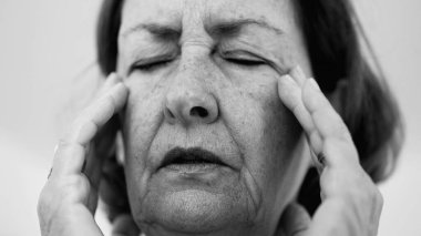Yaşlı bir kadın parmaklarını şakaklarına bastırıyor, gözleri kapalı, şiddetli baş ağrısı ve stres belirtileri gösteriyor. Derin kırışıklıklar ve sıkı yüz ifadesi rahatsızlığı gösteriyor.