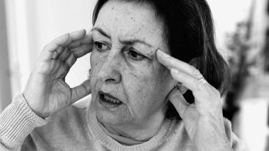 Şiddetli baş ağrısı ile mücadele eden yaşlı bir kadın, gözle görülür konsantrasyonda şakaklara masaj yapıyor. Yüz hatları ve ifadeleri yoğun acıyı, zihinsel gerginliği, duygusal sıkıntıyı yansıtıyor.
