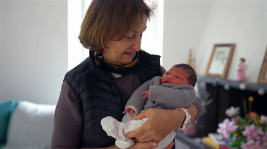 Büyükannem yeni doğmuş bir bebeği gururlu bir gülümsemeyle kucağında tutuyor, sıcak bir yerde duruyor, aile sevgisini ve çoklu-kuşak bağını gösteriyor..