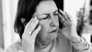 Şiddetli baş ağrısı ile mücadele eden yaşlı bir kadın, gözle görülür konsantrasyonda şakaklara masaj yapıyor. Yüz hatları ve ifadeleri yoğun acıyı, zihinsel gerginliği, duygusal sıkıntıyı yansıtıyor.