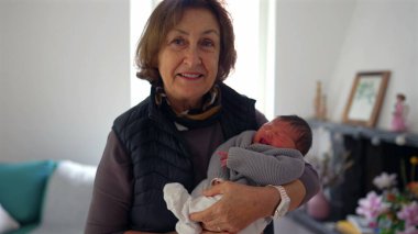 Büyükannem yeni doğmuş bir bebeği gururlu bir gülümsemeyle kucağında tutuyor, sıcak bir yerde duruyor, aile sevgisini ve çoklu-kuşak bağını gösteriyor.