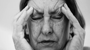 Yaşlı bir kadın parmaklarını şakaklarına bastırıyor, gözleri kapalı, şiddetli baş ağrısı ve stres belirtileri gösteriyor. Derin kırışıklıklar ve sıkı yüz ifadesi rahatsızlığı gösteriyor.
