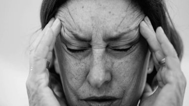 Yaşlı bir kadın parmaklarını şakaklarına bastırıyor, gözleri kapalı, şiddetli baş ağrısı ve stres belirtileri gösteriyor. Derin kırışıklıklar ve sıkı yüz ifadesi rahatsızlığı gösteriyor. SD 480p