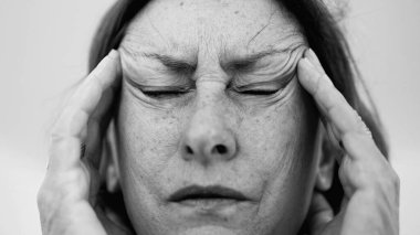 Yaşlı bir kadın parmaklarını şakaklarına bastırıyor, gözleri kapalı, şiddetli baş ağrısı ve stres belirtileri gösteriyor. Derin kırışıklıklar ve sıkı yüz ifadesi rahatsızlığı gösteriyor. SD 480p