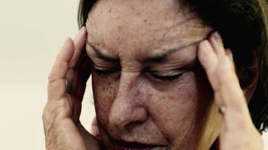 Yaşlı bir kadın şakaklarına sıkıca masaj yapıyor. Acıyı veya stresi azaltmaya odaklanıyor. Derin kırışıklıklar, yoğun yüz ifadesi, görülebilir gerilim şiddetli baş ağrısı gösteriyor. SD 480p