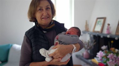 Büyükannem yeni doğmuş bir bebeği gururlu bir gülümsemeyle kucağında tutuyor, sıcak bir yerde duruyor, aile sevgisini ve çoklu-kuşak bağını gösteriyor.