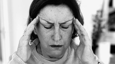 Şiddetli baş ağrısı ile mücadele eden yaşlı bir kadın, gözle görülür konsantrasyonda şakaklara masaj yapıyor. Yüz hatları ve ifadeleri yoğun acıyı, zihinsel gerginliği, duygusal sıkıntıyı yansıtıyor.