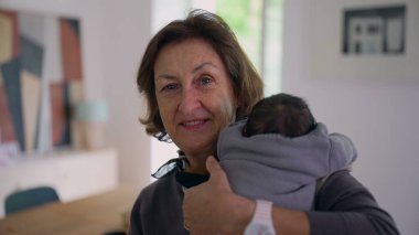 Büyükanne yeni doğmuş bir bebeği omzunda tutuyor, kameraya sıcak bir şekilde gülümsüyor, sıcak bir ev ortamında aile sevgisini ve bağlantısını gösteriyor..