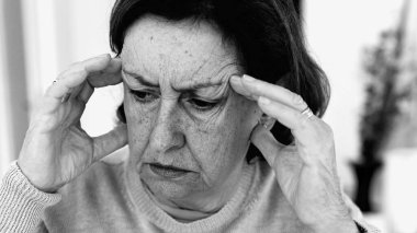 Şiddetli baş ağrısı ile mücadele eden yaşlı bir kadın, gözle görülür konsantrasyonda şakaklara masaj yapıyor. Yüz hatları ve ifadeleri yoğun acıyı, zihinsel gerginliği, duygusal sıkıntıyı yansıtıyor.