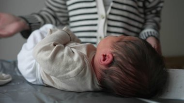 Anne yeni doğmuş bir bebeği nazikçe değişen bir paspasın üzerine yerleştirir, giysiyi özenle ayarlar. Bebek annesine bakıyor ve bir anlık yakınlık ve şefkat gösteriyor. SD 480p.