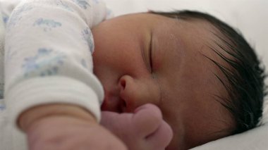Bebek huzur içinde uyurken, yumuşak, narin bir cilt ve gevşek bir ifadeyle yeni doğmuş bir bebeğin yüzünün yakın çekimi. Görüntü, dinlenme ve huzur dolu bir anı yakalar..