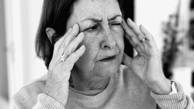 Şiddetli baş ağrısı ile mücadele eden yaşlı bir kadın, gözle görülür konsantrasyonda şakaklara masaj yapıyor. Yüz hatları ve ifadeleri yoğun acıyı, zihinsel gerginliği, duygusal sıkıntıyı yansıtıyor.