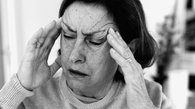 Şiddetli baş ağrısı ile mücadele eden yaşlı bir kadın, gözle görülür konsantrasyonda şakaklara masaj yapıyor. Yüz hatları ve ifadeleri yoğun acıyı, zihinsel gerginliği, duygusal sıkıntıyı yansıtıyor.
