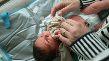 Bebek bakıcı tarafından yumuşak bir paspasın üzerinde nazikçe giydiriliyor. Bebek sakince yukarı bakıyor. Bakıcının elleri elbiseyi özenle ve şefkatle ayarlıyor. SD 480p.