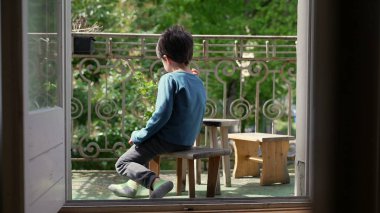 Balkonda tahta bir taburede oturan, yeşilliğe bakan bir çocuğun arka görüntüsü, sakin bir yalnızlık anı, huzurlu bir açık hava ortamında.