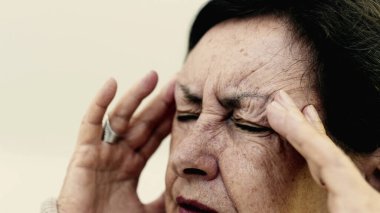 Yaşlı bir kadın şakaklarına sıkıca masaj yapıyor. Acıyı veya stresi azaltmaya odaklanıyor. Derin kırışıklıklar, yoğun yüz ifadesi, görülebilir gerilim şiddetli baş ağrısı gösteriyor. SD 480p
