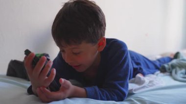 Genç çocuk, evde boş zamanlarında yatakta uzanmış, gülümseyen ve meşgul bir şekilde oyun oynarken ya da içeriği izlerken rahatlamış bir çocukluk anı yaşıyor.