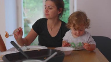 Anne masada oturup yemek yerken, yeni yürümeye başlayan çocuk, evdeki doğal etkileşimi ve kaynaşmayı gösteren otantik aile yemeği sahnesini merakla inceliyor..