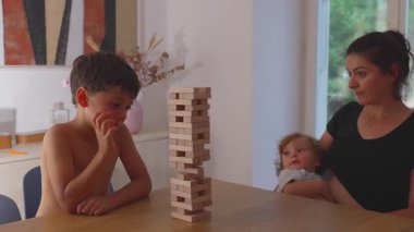 Çocuk, uzun bir istifleme kulesinden tahta bir bloğu kaldırıyor. Annesi kollarındaki bebekle izliyor. Kuleler çökmeden ve bloklar dağılmadan önceki aile dakikalarına odaklanıyor..