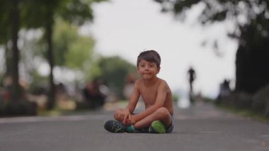 Gülümseyen çocuk, yazın öğleden sonra boyunca park yolunda bacak bacak bacak üstüne atmış, rahat ve memnun, yumuşak yeşillik ve mesafeli insanların bulanık arka planı ile çevrili.