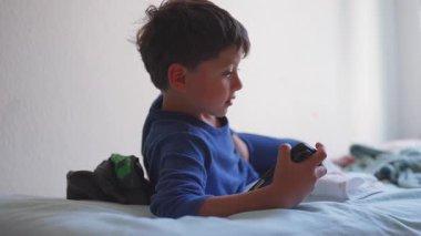 Genç çocuk akıllı telefonu tutarak yatakta uzanıyor, ekranla etkileşime geçerken hafifçe gülümsüyor, evdeki dijital eğlencenin neşeli ve rahat anı.