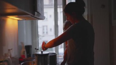Anne Mutfakta Yemek Pişirirken Ocağın yanında Bebek Tutarken Sıcak Akşam Işığıyla Sıcak Ev Ortamı Oluşturuyor.