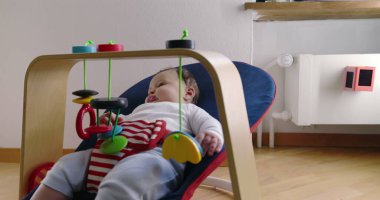 Bebek bir fedainin içinde uzanıyor ve asılı oyuncaklarla oynuyor, rahat bir ev ortamında keşiften ve oynamaktan zevk alıyor, çocukluk yıllarının merak ve masumiyetini vurguluyor.