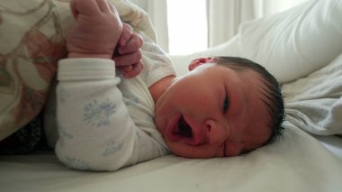 Yeni doğmuş bebek battaniyenin altında hareket ediyor, tuhaf bir ifadeyle yüzünü buruşturuyor, hayatın ilk ayındaki tipik hareketleri ve ifadeleri vurguluyor.